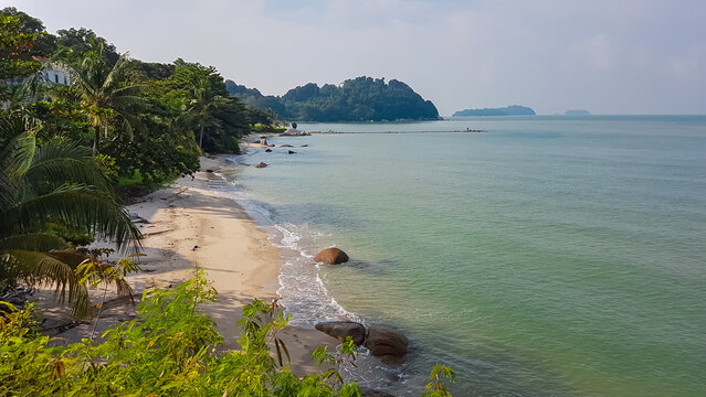 Scenic coastal view with sandy beach curving along calm shoreline of tropical island Pulau Besar near Malacca, Malaysia. Lush green vegetation and trees line coast. Hazy horizon with distant islands