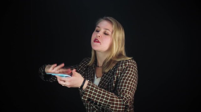 A young smiling joyful woman with blonde hair and a checkered jacket throws pompously dollar bills around on a black background. The concept of success, triumph, wealth and excessive spending of money