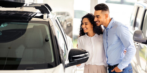 Happy middle-eastern young family choosing new auto in salon, standing by nice white vehicle, looking inside car through window, having conversation, embracing and smiling, copy space