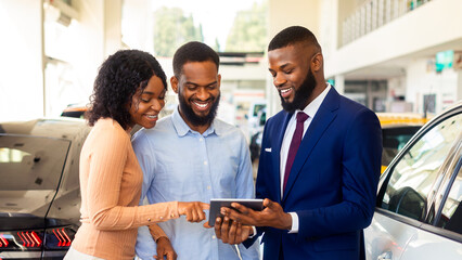 Dealership Center Manager Showing Vehicle Characteristics On Digital Tablet To Young Black Couple, Cheerful African American Spouses Purchasing New Vehicle In Auto Showroom, Closeup Shot