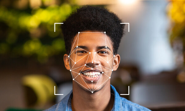 A young man is smiling while facial recognition technology scans his face. This advanced identification method is shown in a modern, well-lit environment with green foliage.