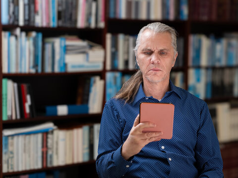 Man with pony tail hair and blue shirt in front of bookshelf is serious while reading on his e-book