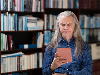 Man with long hair and blue shirt in front of bookshelf is confused while reading on his e-book