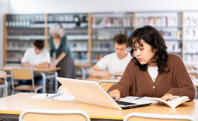 Focused asian teenager female student sitting at library desk with laptop, preparing for exam