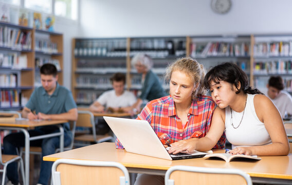 Two fifteen-year-old schoolgirls studying on a laptop and writing abstract in copybooks, doing homework in the school library