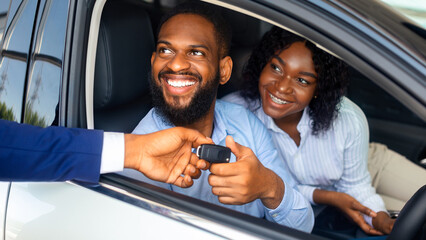 Happy African American Couple Taking Key From Salesman After Buying New Car In Dealership Center,...