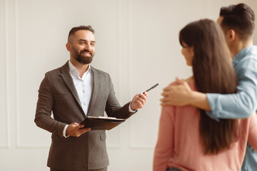Meeting With Realtor Concept. Professional Real Estate Agent In Suit Showing New Home To Young Arab Couple, Millennial Newlyweds Purchasing Or Renting New Flat In Modern House, Choosing First Flat