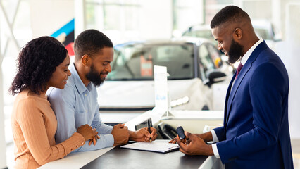 Buying New Car. African American Couple Signing Papers In Dealership Center, Happy Black Spouses...