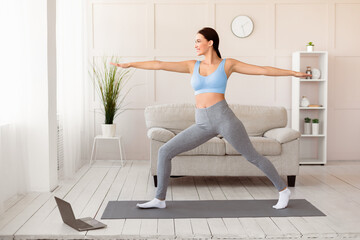 Fototapeta premium Home Leisure. Woman Doing Yoga At Laptop Watching Online Class Standing In Warrior Pose Indoor. Copy Space