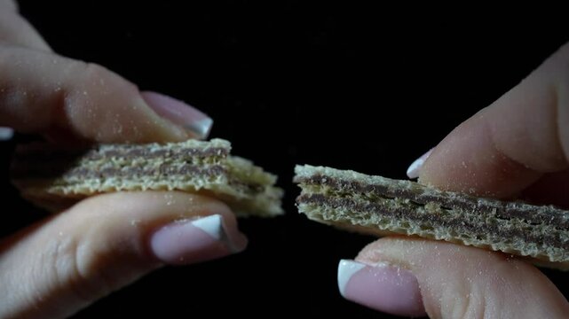 Girl fingers hold a wafer with a chocolate filling, and then break it in half on black background, close up,  slow motion
