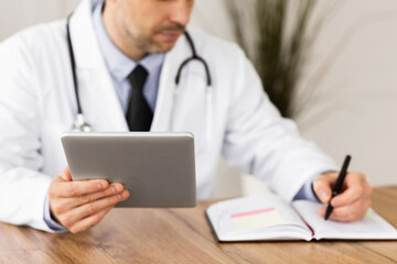 Healthcare, Medical Care And Technology Concept. Male doctor working in office at clinic, writing with pen in notebook, holding and using digital tablet, checking appointment schedule