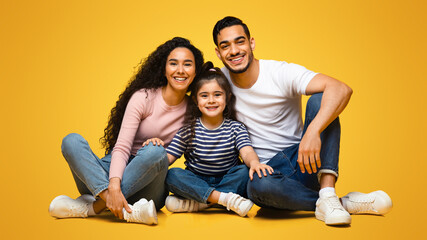 Fototapeta premium Portrait Of Smiling Arab Parents And Little Daughter Posing Together Over Yellow Background, Happy Young Middle Eastern Family Of Three Bonding While Sitting On Floor In Studio, Closeup Shot