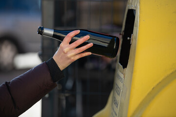 A person is depositing a glass bottle into a yellow recycling bin, engaging in eco-friendly habits....