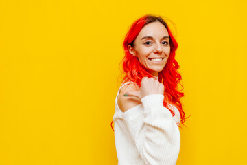 red-haired woman smiling and pointing back on yellow isolated background, girl with colored curly hair showing and advertising copy space behind