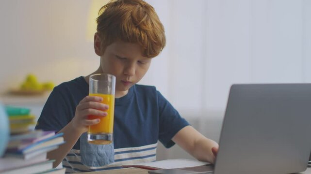 A preteen boy with red hair focuses intently on his laptop while studying at home. He enjoys a glass of orange juice, surrounded by books and learning materials.