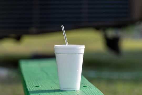 A white drinking cup with a clear plastic straw on a green wooden table. The beverage container holds both cold and hot liquids. The polystyrene a non-biodegradable material.