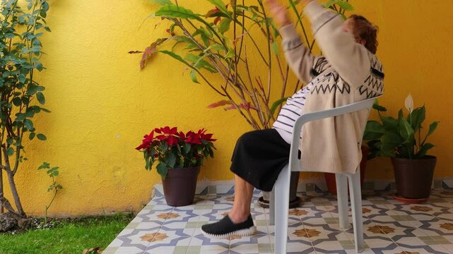 Senior woman exercising legs and abdomen with coordinated up and down leg and arm movements, sitting on chair in garden. Home exercise concept for seniors and healthy lifestyle