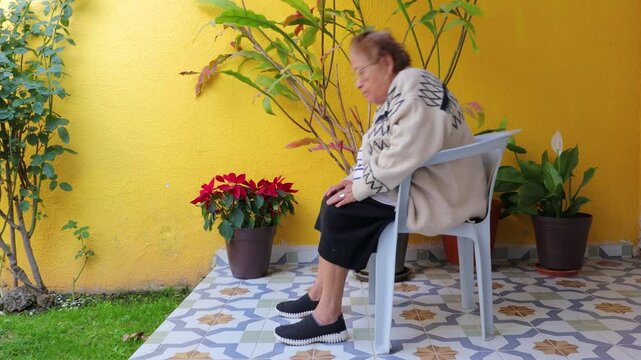 Senior woman exercising her abdomen sitting on chair in garden, leaning back and forth, yellow wall in background. Concept of senior home workout and healthy lifestyle