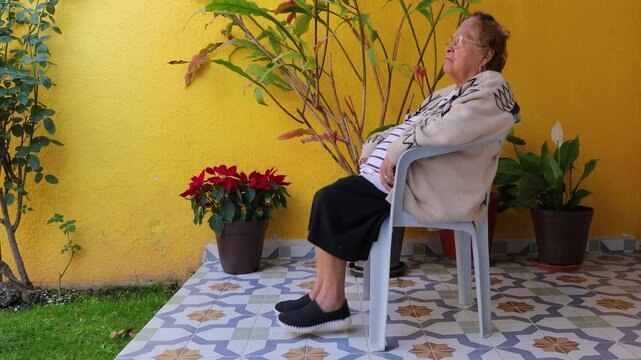 Senior woman exercising legs by moving up and down in coordinated manner, working out while sitting on chair in the garden. Home exercise concept for seniors and healthy lifestyle
