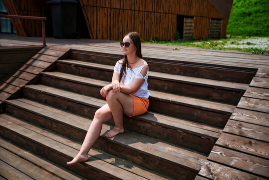 A barefoot Russian woman in her 40s sits on wooden stairs in summer, wearing orange shorts and a white blouse. She enjoys the sunny weather with a relaxed expression - Powered by Adobe