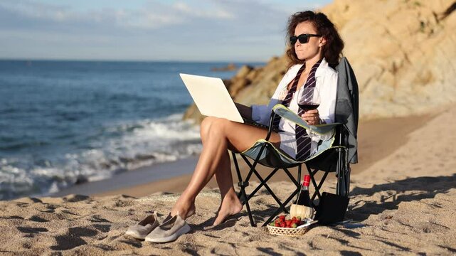 Seductive caucasian businesswoman wearing only white suit jacket, with naked breast, sitting on blanket and posing near shoreline during picnic with wine and fruits. 