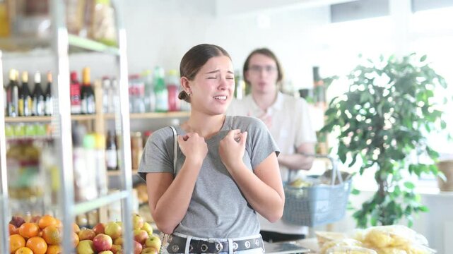 In shop guy stands behind wife, hurling abuse and indignation at her. In trading floor, sullen man stands with shop basket, swears with wife and listens to womans accusations.