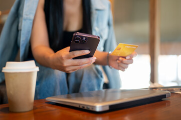 A close-up of a woman sits in a coffee shop, holding her phone and a credit card, shopping online.