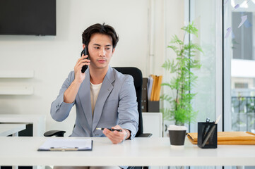 A confident and serious Asian businessman is talking on the phone while working in his office.
