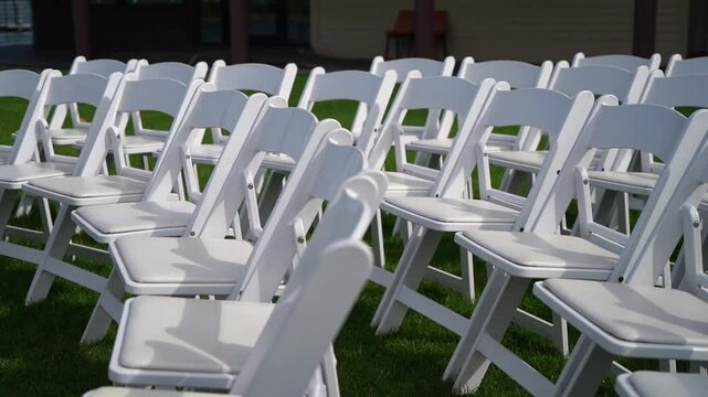 Rows of white folding chairs are arranged on a lush green grass lawn, perfect for an outdoor event such as a wedding or ceremony. The seating is prepared for guests to sit comfortably