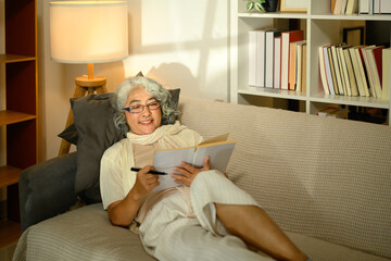 Happy elderly woman with gray hair sitting on sofa and writing in notebook with a smile