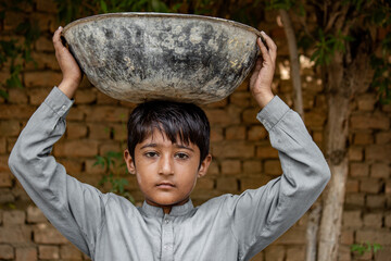 Young boy in traditional dress carrying a large bowl on his head, symbolizing child labor and resilience in rural life.
