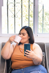 Plus size latina woman sitting on sofa drinking coffee and looking at smartphone