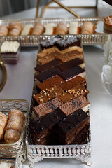 table with various desserts. In the foreground are three metal trays. The first tray on the left contains a variety of cakes, including chocolate frosting and nuts.