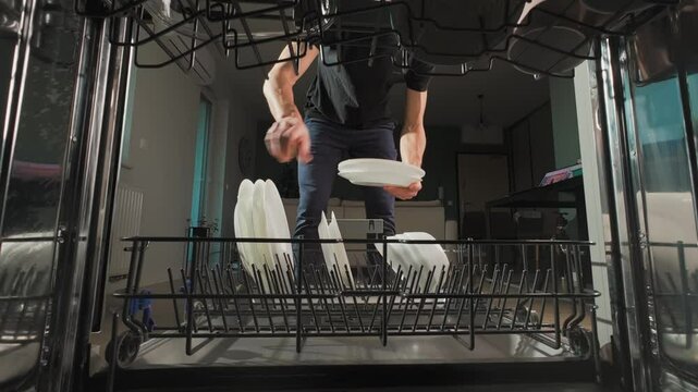 Man unloading clean white plates from the bottom rack of dishwasher in kitchen. Person performing everyday household chores, using modern home appliance