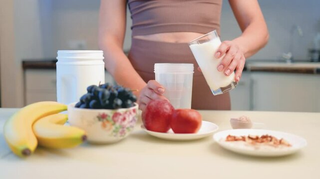 Athletic woman in sportswear prepares protein drink in a kitchen, pouring milk and putting a portion of whey protein powder into a shaker on a table, slow motion