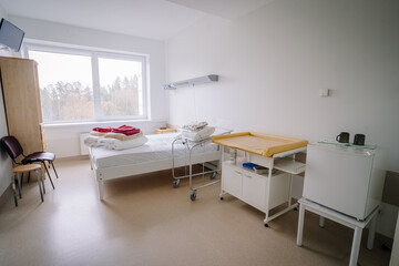 Bright hospital room with a single bed, changing table, storage units, chairs, and a large window overlooking trees, creating a clean and calm atmosphere.