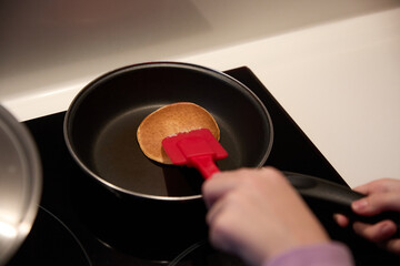 Cooking a pancake in a frying pan on the induction cooker at home with a red plastic spatula
