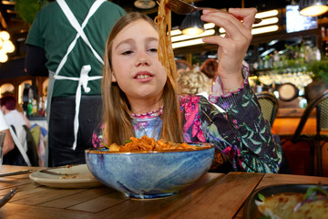 10 year old girl ready to eat holding with fork noodles of pad thai with chicken from a blue bowl on a wooden table in a restaurant
