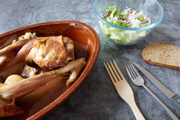 Roasted lamb legs on a clay platter with fork, knife, green lettuce and onion salad and slice of bread on a marble texture table