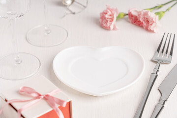 Empty ceramic white plate of heart shape served on wooden table with pink carnation flowers, gift box with ribbon, wine glass, knife and fork prepared for celebration of romantic valentines day
