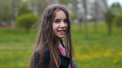 Naklejka premium Teenage girl with long brown hair smiling, wearing leather jacket, standing in lush green park during sunny spring afternoon