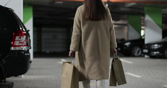 Caucasian woman shopper walks through mall parking lot holds cardboard bag in hand open trunk of car.