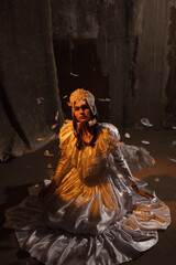 Beautiful young brunette dressed as an angel kneels in a dimly lit room, draped in an ornate white...