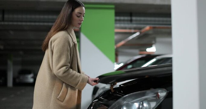 Woman stranded near dysfunctional car in the parking lot of shopping center with open hood, looking at broken engine.