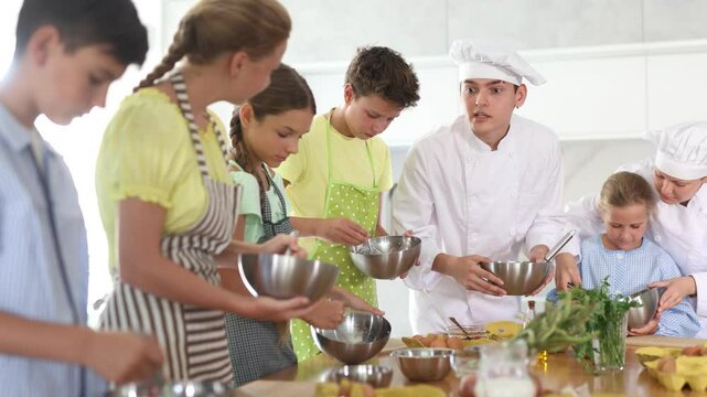 Positive woman and guy, professional chefs in white uniform conducting culinary masterclass together, imparting cooking skills to group of interested tween boys and girls