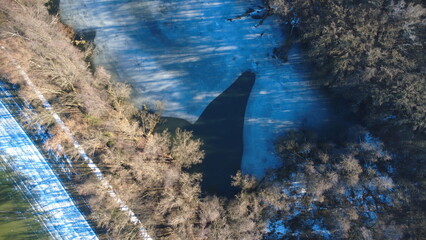 Aerial view of a frozen lake in a forest during the spring frost season. Surrounding the lake are...