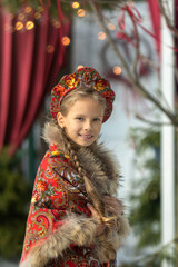 A blonde girl in a national Russian costume on the Maslenitsa holiday. A child in a traditional costume with fur trim and a headdress against a festive backdrop. Winter scene with a beautiful girl