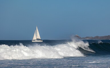 Sailboat navigates the ocean with waves crashing under a clear blue sky, near a distant coastline