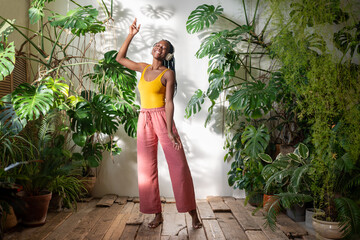 Serene African American young woman relaxing to songs amidst lush houseplants, savour moment. Carefree black woman dancing barefoot in headphones, immersed in music on wooden floor experience pure joy