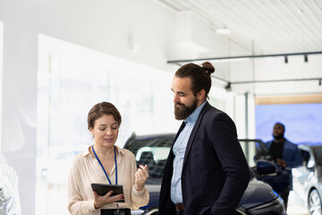 Car dealership salesman reading text on tablet to customers, providing information about available models, features and pricing options. Agent negotiating vehicle prices with clients